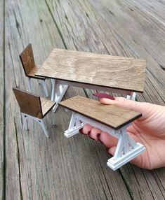 someone is holding a miniature school desk and chair on a wooden table with one hand