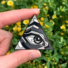 a hand holding a triangle shaped object with an eye in the center and yellow flowers behind it