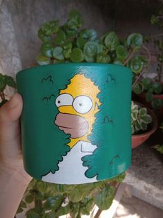 a person holding up a green cup with the simpsons on it's face in front of a potted plant