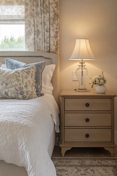 a white bed sitting under a window next to a night stand and lamp on top of a dresser