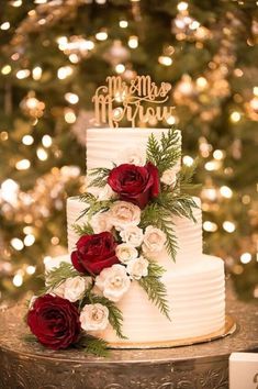 a white wedding cake with red and white flowers on top is sitting in front of a christmas tree