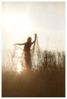 a woman standing in tall grass with her arms outstretched and the sun shining behind her