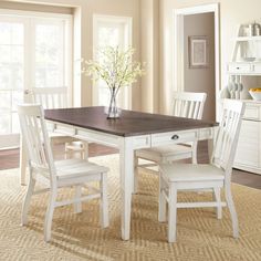 a dining room table with white chairs and a vase on top of it in front of windows