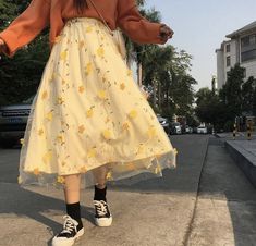 a woman walking down the street in a yellow dress and black tennis shoes with an orange sweater over her shoulders