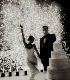 a bride and groom standing next to a wedding cake with fireworks in the background at night