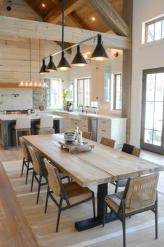 a dining room table with chairs and lights hanging from it's ceiling in front of an open kitchen