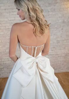 a woman in a white wedding dress with a big bow on her back, standing against a brick wall