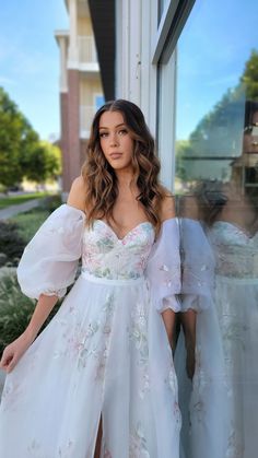 a woman standing in front of a window wearing a white dress with flowers on it