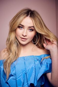 a woman with long blonde hair wearing a blue top and posing for a photo in front of a pink wall