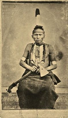 an old black and white photo of a native american man sitting on a rock with a knife in his hand