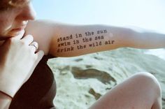 a woman sitting on the beach with her arm tattoo saying, stand in the sun swim in the sea drink the wild air