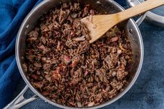 a wooden spoon in a pot filled with meat and vegetables on a blue cloth next to two silver pans