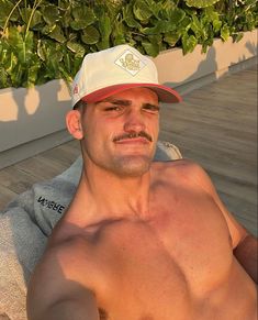 a shirtless man wearing a hat and sitting on a wooden deck next to plants