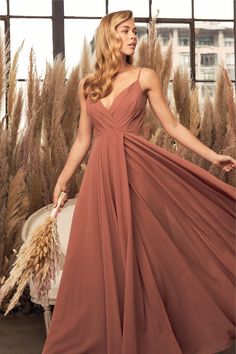 a woman in a long brown dress standing next to some tall grass and looking off into the distance