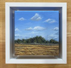 a painting of a field with trees and clouds in the sky, on a wooden surface