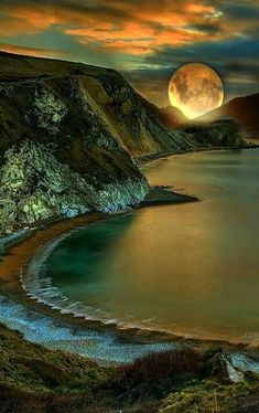 a full moon setting over a body of water with hills in the background and grass on the shore