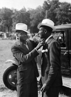 two men standing next to each other in front of an old car with hats on