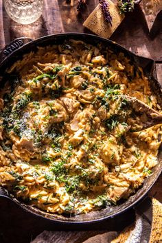 a skillet filled with pasta and vegetables on top of a wooden table next to wine glasses