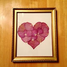 a heart made out of pink flowers in a gold frame on a wooden table top