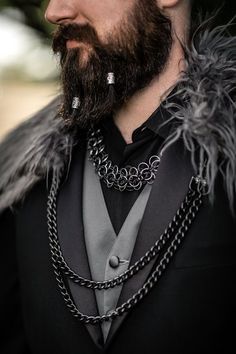 a bearded man in a black suit with chains on his neck and collar, wearing an elaborate necklace