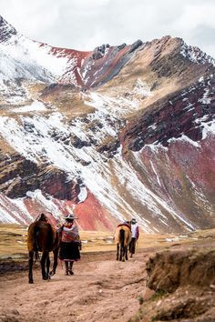 three horses walking down a dirt road in front of snow covered mountain peaks, with two people on the back