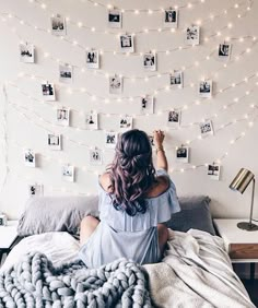 a woman sitting on top of a bed in front of a wall with pictures and string lights