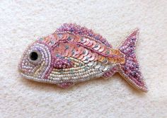 a pink and gold fish brooch sitting on top of a white blanket next to a black eye