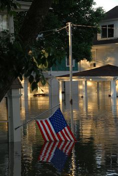 an american flag is in the middle of a flooded street with houses and trees on either side