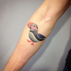 a small colorful bird tattoo on the left arm and wrist, with an arrow in it's beak