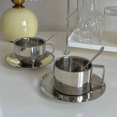 two silver pots and saucers sitting on a white counter top next to each other