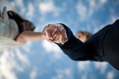 two people holding hands in the air with clouds and blue sky behind them, one pointing at the other