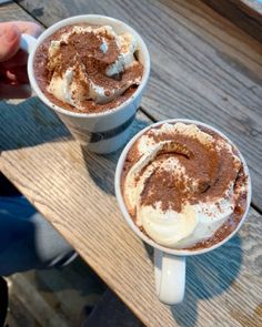 two cups of hot chocolate with whipped cream and cinnamon on the top, sitting on a wooden table