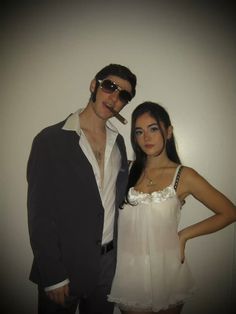 a man and woman pose for a photo in front of a white wall wearing sunglasses