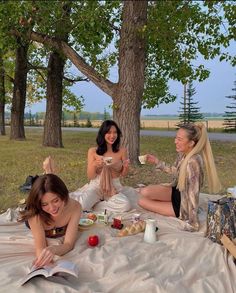 three beautiful women sitting on top of a blanket in the park eating food and drinking