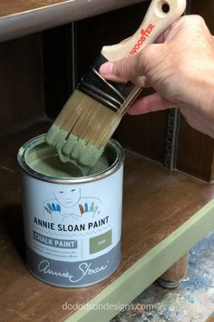 a person holding a paint brush over a can of white paint on top of a wooden shelf