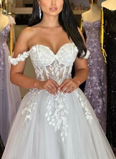 a woman standing in front of dresses on mannequins wearing an off the shoulder wedding dress