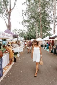 a woman in a white dress and hat walking down the street at an outdoor market