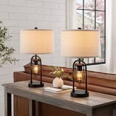 two lamps sitting on top of a wooden table next to a brown couch and window