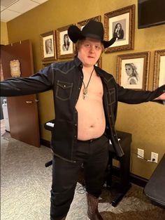 a man with no shirt and cowboy hat standing in an office wearing black pants and boots