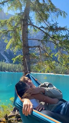 a woman laying in a hammock next to a tree on the shore of a lake