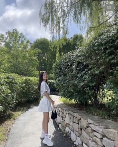 a woman standing on the side of a road next to a stone wall and trees