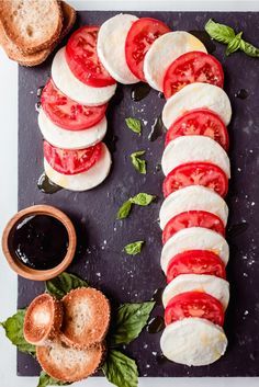 sliced tomatoes, mozzarella and basil on a black cutting board with bread in the background