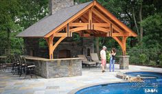 two people standing in front of an outdoor kitchen next to a pool with a fireplace