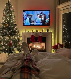 a person laying in bed with their feet on the pillow watching tv next to a christmas tree