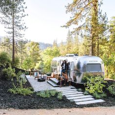 an rv is parked in the woods with people standing outside and sitting on steps leading up to it