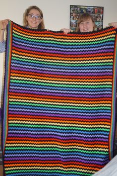 two women holding up a multicolored crocheted blanket