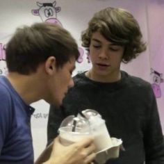 two young men standing next to each other looking at something in a paper bag on top of a table