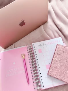 a pink notebook with a gold pen next to an apple laptop on a white bed