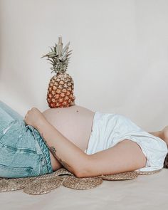 a pregnant woman laying on top of a bed next to a pineapple sitting on her stomach