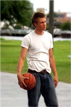 a young man holding a basketball in his hand while standing on the sidewalk with grass and trees behind him